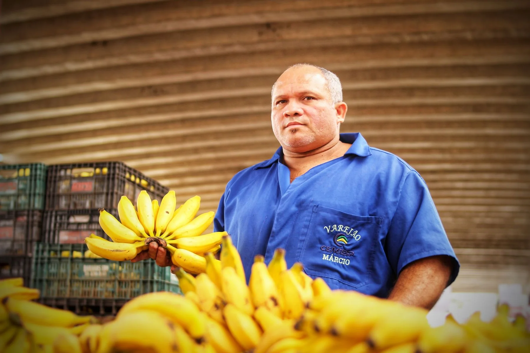 Feirante de hortifrúti segurando um cacho de bananas em sua banca